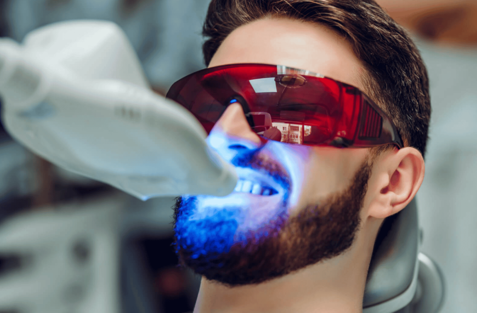 A man in a dentist's office wearing specialty red wraparound glasses to protect his eyes from UV rays being projected from a device in front of his mouth and on to his teeth during a teeth whitening procedure.