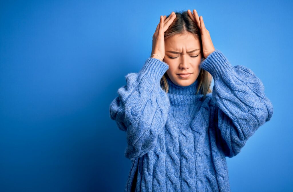 A woman in a blue sweater places both of her hands on her head due to headache pain.